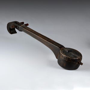 May include: A dark brown stringed musical instrument with a rounded body and a long neck. The instrument has a curved headstock and appears to be made of wood. The body has a textured, patterned surface.
