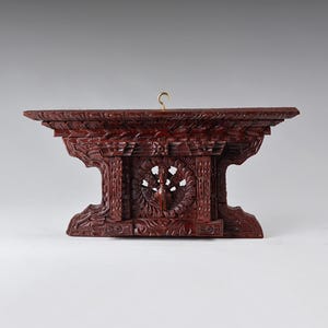 May include: A dark brown, hand-carved wooden wall shelf featuring intricate floral and geometric patterns. The central panel displays a circular motif with a bird figure. A small, gold-coloured metal hook is attached to the top centre edge.