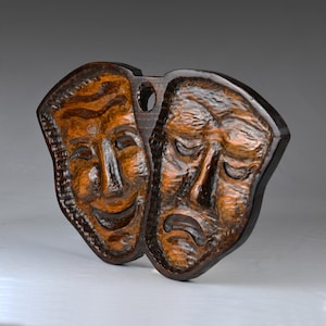 May include: A wooden carving of two theatrical masks, one smiling and one frowning. The masks are brown and have a textured surface.