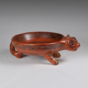 May include: A reddish-brown ceramic bowl shaped like a quadruped animal. The bowl has a rounded form with a decorative rim and is supported by four legs. The animal's head is detailed with facial features and the bowl is decorated with black patterns.
