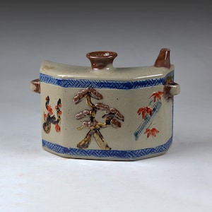 May include: A beige ceramic sake bottle with a blue geometric border and brown accents. The bottle features hand-painted images of a tree, bamboo, and flowers. It has a spout and a handle on each side.