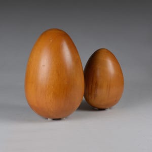 Puede incluir: Dos objetos decorativos de madera con forma de huevo. El huevo más grande está en primer plano, con otro más pequeño detrás. Ambos son de color marrón claro cálido con un acabado liso. Los huevos tienen forma redondeada y se apoyan en pequeñas bases blancas.