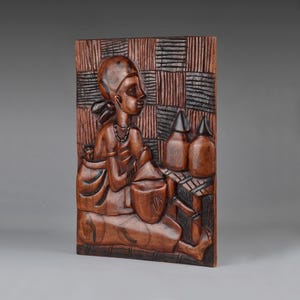 May include: A carved wooden relief showing a person seated with a child on their back. The person wears a head covering and jewellery. The background has a checkered pattern. Several bottles are visible.