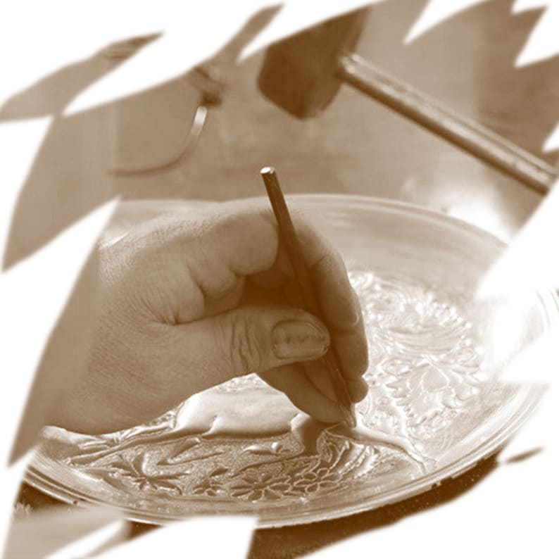 May include: A hand using a tool to create a design on a metal plate. The plate has a detailed pattern and is being worked on by a craftsperson.