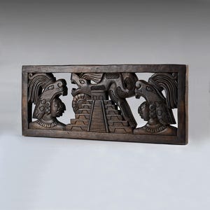 May include: A rectangular dark brown wooden wall carving featuring a central stepped pyramid flanked by two figures wearing ornate headdresses. A large serpent figure is positioned above the pyramid, integrated into the relief design.