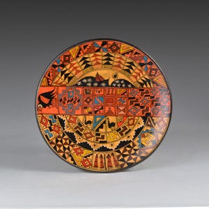 以下が含まれることがあります： 黒い縁と鮮やかな幾何学模様が特徴の丸い装飾皿。皿の中央には鳥のモチーフがあり、オレンジ、赤、黄色、青、黒の模様で囲まれています。デザインには幾何学的な形と様式化された動物の姿が含まれています。