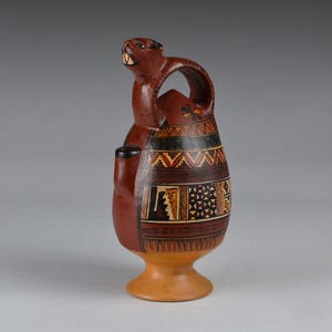 May include: A brown ceramic vessel with a beaver-like figure on top and a handle. The vessel features geometric patterns in black, tan, and white. The base is a lighter tan color.