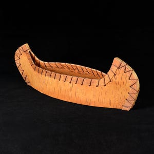 Könnte beinhalten: Ein dekoratives Modellkanu aus hellbrauner Birkenrinde mit natürlicher Holzmaserung. Die Struktur wird durch eine dünne, dunkelbraune Kordelschnürung entlang der oberen Kanten sowie an Bug und Heck zusammengehalten.
