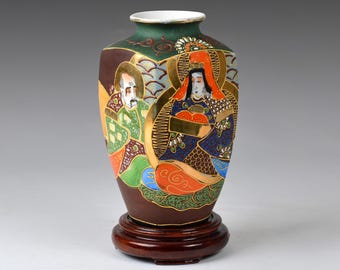 May include: A brown ceramic vase with a green, gold and blue painted design of two figures in traditional Japanese clothing. The vase is on a wooden base.