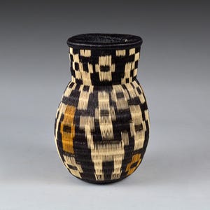 Puede incluir: Una cesta tejida con un cuerpo bulboso y un cuello estrecho. La cesta presenta un patrón geométrico de fibras negras y de color natural, con acentos dorados. El borde es negro sólido.