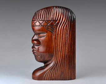 May include: A carved wooden sculpture of a human head in profile. The sculpture is a rich, reddish-brown color and features detailed carvings of hair, facial features, and a headband. The style suggests a tribal or cultural artifact.