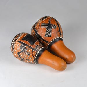 Pode incluir: Duas maracas de cabaça com intrincados desenhos gravados em preto e branco. As cabaças têm uma cor bronzeada natural com uma faixa preta perto da pega. Os desenhos incluem padrões geométricos e figuras estilizadas, sugerindo um tema cultural ou tribal.