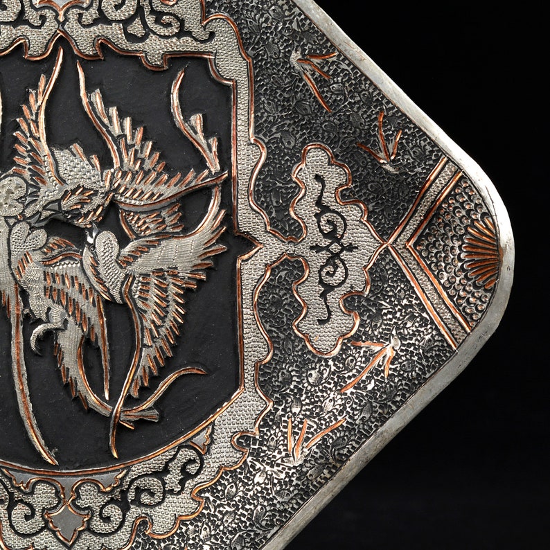 May include: A silver and copper tray with an intricate design of birds and floral motifs. The tray has a black background and is decorated with a raised, textured pattern.