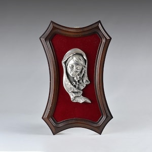 May include: A wall plaque featuring a silver-toned metal relief of a person wearing a head covering and holding a small flower. The relief is mounted on a deep red velvet backing and encased within a dark brown wooden frame with curved edges.