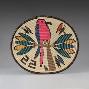 May include: A round woven basket featuring a central pink and blue parrot perched on a brown branch. The design includes symmetrical teal and yellow fan-like patterns on both sides, with black geometric accents on a cream-colored background.
