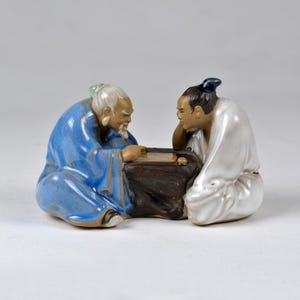 Puede incluir: Dos figuritas de cerámica de hombres jugando a un juego de mesa. Los hombres visten ropa tradicional china. El hombre de la izquierda lleva una túnica azul y el hombre de la derecha lleva una túnica blanca. El juego de mesa está sobre una mesa marrón.