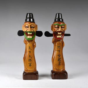 Puede incluir: Dos figuras de madera tallada con caras sonrientes, sombreros negros y accesorios laterales negros. Las figuras tienen detalles pintados en verde, rojo y blanco. Cada figura tiene caracteres chinos tallados en el frente. Las figuras están montadas sobre bases marrones oscuras.