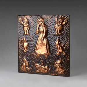 Könnte beinhalten: Kupferfarbene Metallreliefskulptur mit einer zentralen Figur einer Frau in einem langen Kleid, umgeben von sieben Zwergen. Der Hintergrund ist mit kleinen, gleichmäßig verteilten Punkten strukturiert. Die Zwerge sind in verschiedenen Posen.