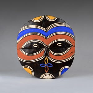 May include: A round wooden mask with a stylized human face. The mask is painted in black, red, white, blue, and yellow. The eyes are large and black, and the mouth is a simple line. The mask is decorated with geometric patterns.