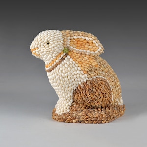 May include: A seated rabbit figurine made from small seashells in shades of white, tan, and brown. The ears are upright, and the figurine has a detailed texture. The colour palette is natural and earthy, suitable for home decor.