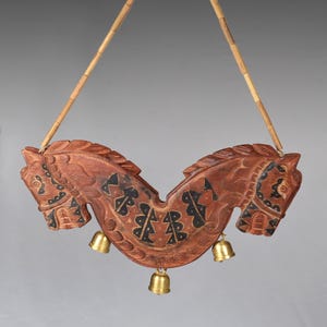 May include: A wooden carving of two horse heads facing outward, with black and brown geometric designs. The carving is attached to a bamboo string and has four small gold bells hanging from the bottom.