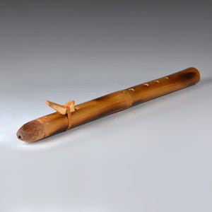 Puede incluir: Una flauta de madera artesanal de color marrón claro. El instrumento presenta una boquilla redondeada y varios agujeros para los dedos a lo largo del cuerpo. Una pequeña pieza de madera de color claro está sujeta con una fina banda marrón.
