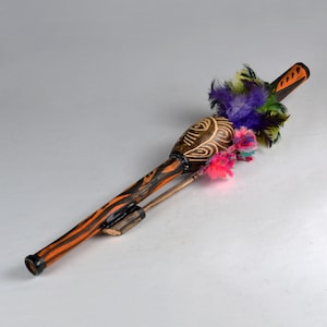 Könnte beinhalten: Ein langes, zylindrisches Holzinstrument mit einem schwarz-orange gestreiften Muster. Der obere Teil ist mit einem Büschel aus violetten, grünen und gelben Federn sowie leuchtend pinken und mehrfarbigen Faserbüscheln verziert.