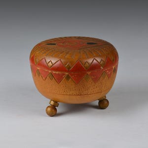 May include: A small, round wooden box with a lid, supported by three small ball feet. The box is light brown with red and gold geometric patterns around the rim and on the lid. The lid has a central circular design.