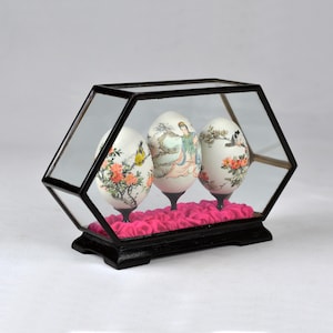 May include: Three hand-painted eggs displayed in a glass hexagonal case with a black wooden base. The eggs are decorated with traditional Chinese scenes and are displayed on pink fabric.