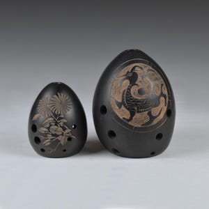 Pode incluir: Duas ocarinas de cerâmica preta. Uma é menor com um desenho floral, a outra é maior com um desenho de pássaro. Ambas têm vários orifícios para os dedos e têm formato de ovo. São instrumentos musicais.