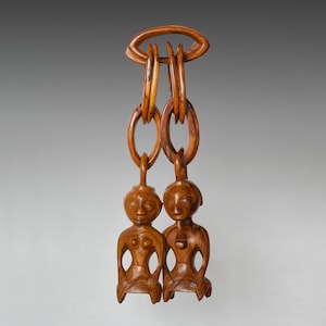 May include: A wooden pendant with two carved figures of people hanging from a chain of interlocking rings. The figures are seated and have detailed facial features.