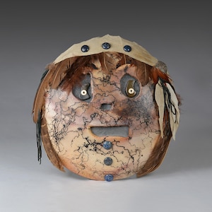 May include: A handcrafted mask with a round face shape, featuring a tan and brown color scheme with intricate black line details. The mask has feather and bead embellishments, with a neutral background.