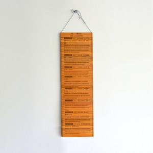 May include: A rectangular, wooden wall hanging with the title "THE ANALECTS" at the top. The text is in both English and Chinese. The wooden slats are a warm, orange-brown color, and the piece is suspended by a thin, black string.