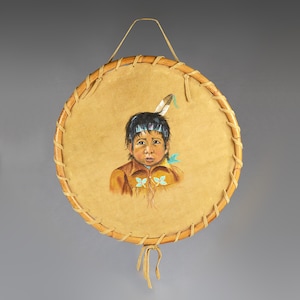 Könnte beinhalten: Runde, handbemalte indianische Trommel mit einem Kinderporträt. Die Trommel hat eine hellbraune Lederfläche mit einem gemalten Bild eines Kindes, das ein braunes Hemd und eine Feder im Haar trägt. Sie ist mit einem hellbraunen Holzrahmen und einem Lederaufhänger versehen.