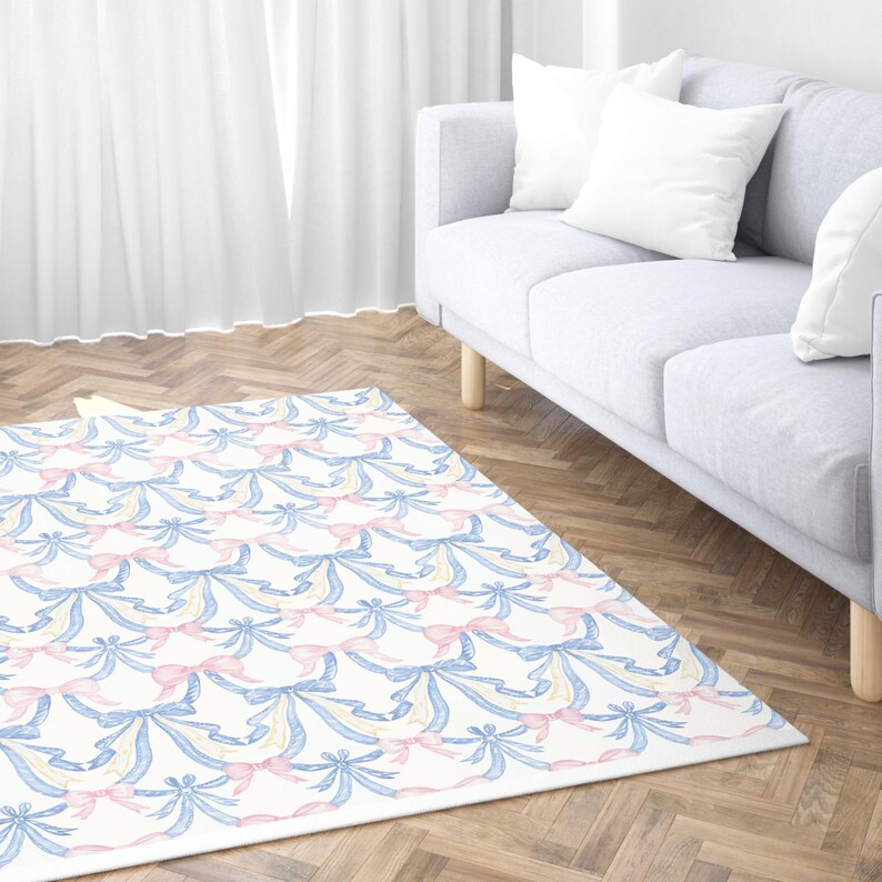 May include: A rectangular area rug with a repeating pattern of blue, pink, and yellow bows on a white background. The rug is placed on a hardwood floor in front of a light gray sofa with white pillows. The room has a bright, airy feel.
