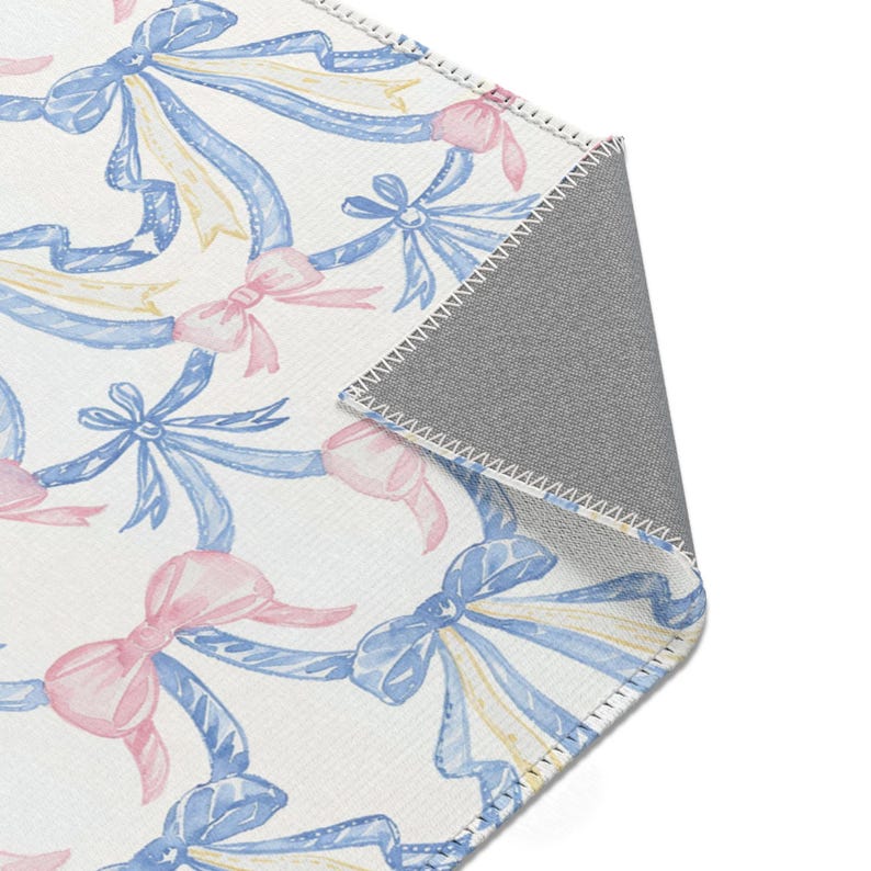 May include: A rectangular rug with a repeating pattern of watercolor bows and ribbons. The bows are in shades of pink and blue, with yellow accents, set against a white background. The rug has a gray backing.