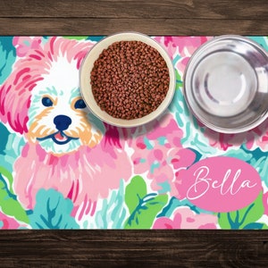 Op de afbeelding: Een roze en groene bloemen hondenvoermat met een witte hondenillustratie en de naam "Bella" in een roze ovaal.