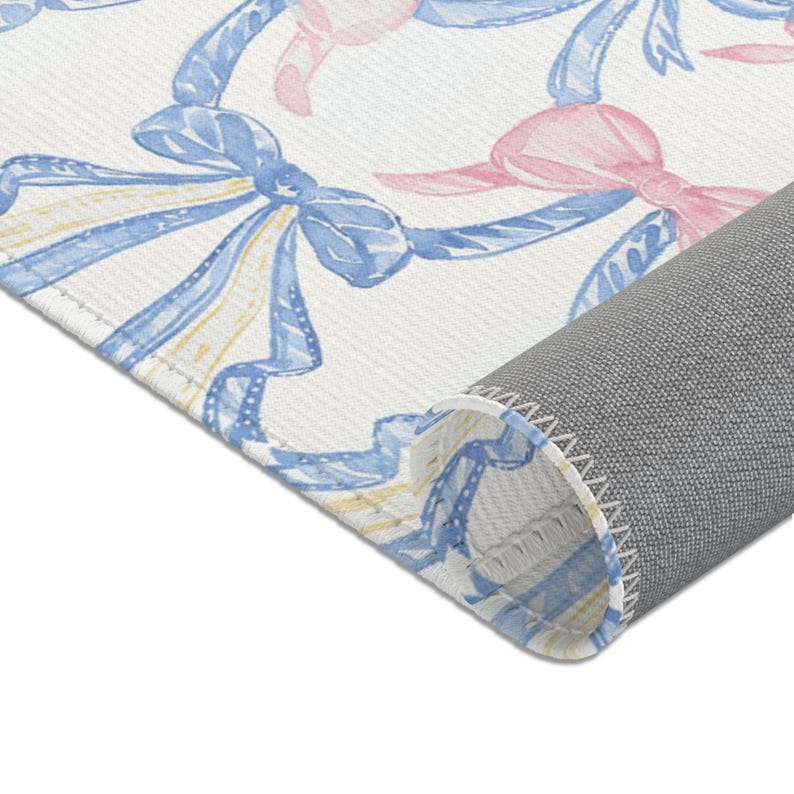 May include: A rolled-up rug with a repeating pattern of watercolor bows in blue, pink, and yellow on a white background. The rug's underside is gray.