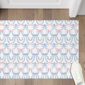 May include: A rectangular doormat with a repeating pattern of blue and pink bows on a white background. The bows are arranged in a symmetrical design. White sneakers are visible at the bottom right.