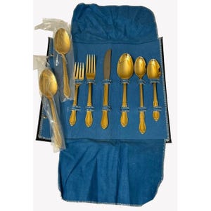 May include: A gold-colored flatware set, including spoons, forks, and a knife, is displayed on a blue fabric. The utensils are secured with blue and white rope-like ties. Some pieces are still in their original packaging.