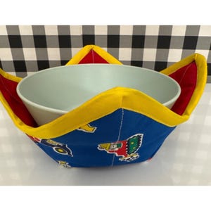 May include: A blue bowl cosy with a colourful pattern of vehicles and toys. The cosy has red and yellow trim and is designed to hold a bowl. The bowl is light blue and sits inside the cosy.