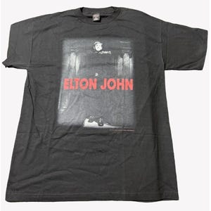 May include: Black t-shirt with a faded image of Elton John in a black suit and white shirt. The text "Elton John" is printed in red letters on the front of the shirt.