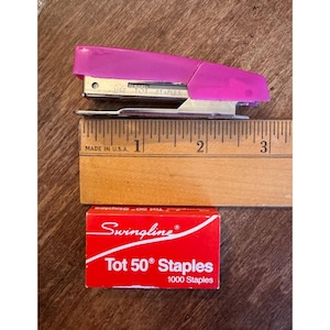 May include: A magenta stapler with silver metal components, a wooden ruler, and a red box of Swingline Tot 50° Staples. The ruler is marked in inches and labeled "MADE IN U.S.A."