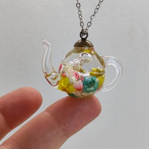 Puede incluir: Un collar con una tetera de vidrio transparente con una cadena plateada. La tetera está llena de flores secas de colores y una tapa dorada.