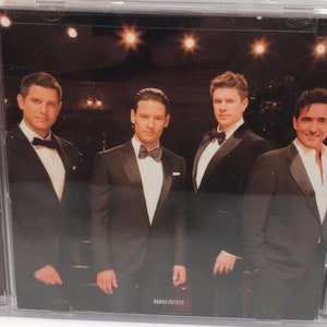 May include: Four men in black suits and bow ties pose for a photo. They are all smiling and looking at the camera. The background is a dark stage with spotlights.