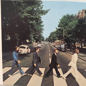 May include: The Beatles Abbey Road album cover features four men walking across a crosswalk in a black and white striped pattern. The men are wearing suits and are walking in the same direction. The background is a street with trees and buildings.