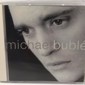 May include: A black and white photo of Michael Bublé on a CD cover. The photo shows a close-up of his face, with his eyes looking down and to the right. The text "michael bublé" is printed in white letters on the bottom of the image.
