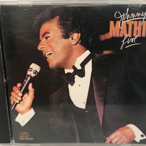 May include: A CD case with a black background and a photo of Johnny Mathis singing into a microphone. The text "Johnny Mathis Live" is printed in white and orange on the case.
