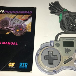 May include: A white and gray Super Nintendo Entertainment System (SNES) controller with a clear plastic shell. The controller has a digital display, buttons, and a directional pad. The controller is connected to a gray cable with a connector for the SNES console. The image also includes a user's manual for the controller.