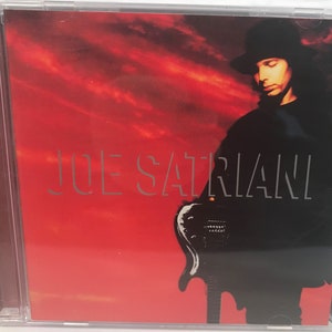 May include: A red and black CD case with a photo of Joe Satriani holding a guitar. The background is a red sunset.